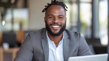 A smiling black man with dreadlocks sitting at a table with a laptop photo