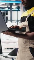 Vertical Black engineer in photovoltaics factory using laptop to diagnose malfunctioning systems, debugging. Solar panels manufacturing plant worker reduces downtime, troubleshooting. Camera B. video