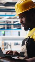 Vertical Male engineer in solar panel factory reviews photovoltaic system performance on laptop. Specialist analyzing production line on notebook to optimize efficiency, troubleshooting. Camera B. video