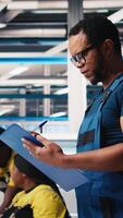 Vertical Black engineer in photovoltaics factory reviews performance of solar panels assembly lines, checking paperwork for finetuning. Supervisor inspecting solar cell machinery. Camera B. video