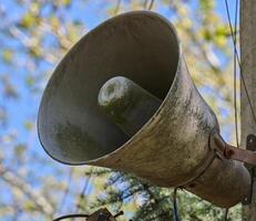 An old, dirty, grey loudspeaker is mounted on a pole. In the background, there is a blurred image of the sky and tree branches. photo