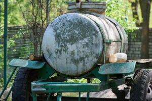 An old metal tank on a trailer, covered with traces of rust and time, stands against the background of green trees and a chain-link fence. photo