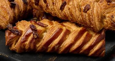 Brown maple pecan braid pastries are arranged on a dark, textured surface. Braided dough is flaky and topped with pecans. photo