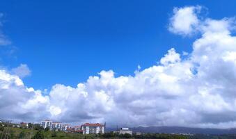 Serene landscape with fluffy white clouds against a bright blue sky. Buildings peek through trees on a distant hill, ideal for background, nature, or travel concepts. photo