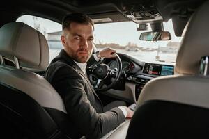 Looking behind at the camera. Man in black suit is sitting in the car,,, driving photo