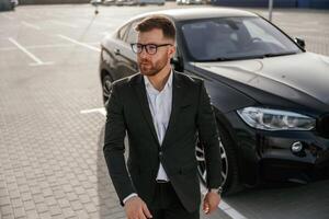 Handsome smart guy in glasses. Businessman in suit is near his black car outdoors.. photo