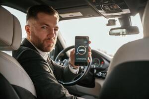 Monitoring charging, process, app, in, smartphone. Man in black suit is sitting in the car, driving photo