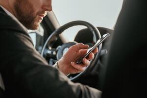 Surfing the internet by using smartphone.,, Man in black suit is sitting in the car, driving photo
