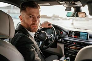 Looking behind at the camera.,, Man ,in ,black suit is sitting in the car, driving photo