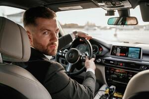 Looking, behind at the camera. Man in black suit is sitting in the car, driving photo