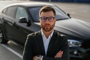 In glasses,, front ,view. Businessman in suit is near his black car outdoors photo