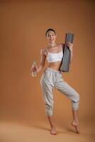 Looking ,forward, holding ,bottle of water and mat. Beautiful fitness woman is in the studio photo