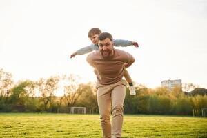 Wellbeing, kid ,is, on, the shoulders or back. Happy father with son are having fun on the field at summertime photo