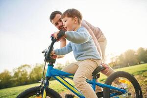 Riding, on ,,the blue bicycle. Happy father, with son are having fun on the field at summertime photo