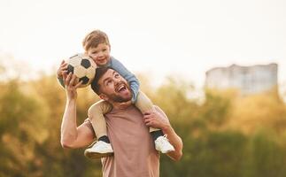On, shoulders,,, with soccer ball. Happy father with son are having fun on the field at summertime photo