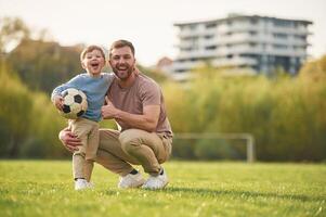 Sitting, holding, soccer ,,ball. Happy father with son are having fun on the field at summertime photo
