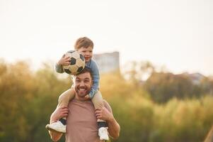 On shoulders, with soccer ball. Happy,, father with son are having fun on the field at summertime photo