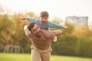 Wellbeing, kid is on the shoulders or back... Happy father with son are having fun on the field at summertime photo