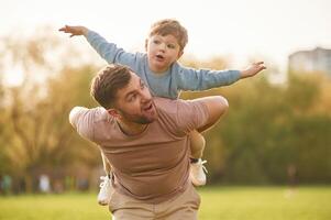 Sitting on man's shoulders... Happy father with son are having fun on the field at summertime photo