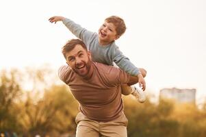 Conception of imagination, sitting on shoulders and flying. Happy father with son are having fun on the field at summertime photo