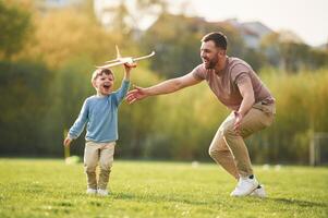 In casual clothes. Running with toy plane. Happy father with son are having fun on the field at summertime photo