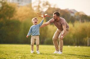 In casual ,,clothes. Running with toy ,plane. Happy father with son are having fun on the field at summertime photo