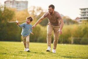 In casual ,clothes. Running with toy plane. Happy father with son are having fun on the field at summertime photo
