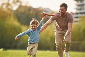 Flying high, holding toy,, plane, running. Happy father with son are having fun on the field at summertime photo