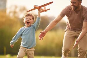 Flying, high, ,,holding toy plane, running. Happy father with son are having fun on the field at summertime photo