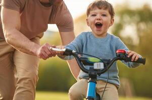 Close up view, excited boy is riding bicycle. Happy father with son are having fun on the field at summertime photo