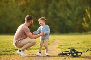 Boy is crying, fell of the bicycle. Happy father with son are having fun on the field at summertime photo