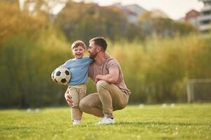 Sitting, holding soccer ball. Happy father, with son are having fun on the field at summertime photo