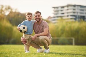 Sitting, holding soccer ball.. Happy father with son are having fun on the field at summertime photo