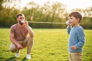 Talking by, using paper cup telephone. Happy father with son are having fun on the field at summertime photo
