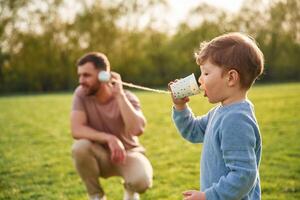 Talking, by,, using paper cup telephone. Happy father with son are having fun on the field at summertime photo