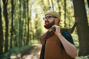 Holding an axe. Bearded man is in the forest at daytime photo