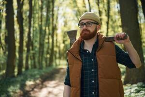 Holding an axe. Bearded man is in the ,forest at daytime photo
