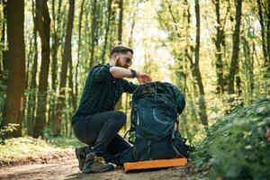 Looking for stuff, in the backpack, sitting. Bearded man is in the forest at daytime photo