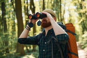 Using, binoculars. Bearded man ,,is in the forest at daytime photo
