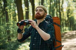 Using ,binoculars. Bearded man is in the forest at daytime photo