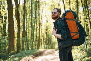 In glasses and with backpack. ..Bearded man is in the forest at daytime photo