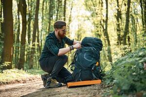 Looking, for stuff in the backpack, sitting. Bearded man is in the forest at daytime photo