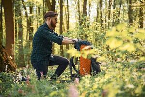 Looking for, stuff ,in the backpack, sitting. Bearded man is in the forest at daytime photo