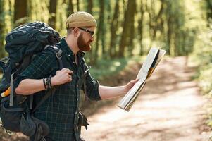 Focused, on reading the map. Bearded man is in the forest at daytime photo