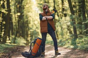 Yellow, axe in ,hand, backpack on the ground. Bearded man is in the forest at daytime photo
