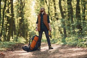 Yellow axe ,in, hand, backpack on the ground. Bearded man is in the forest at daytime photo
