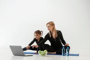 Laptop, apples and bottles of water on the floor. Mother and little daughter are in fitness clothes on yoga mat photo