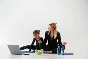 Laptop, apples ,and bottles of water on the floor. Mother and little daughter are in fitness clothes on yoga mat photo