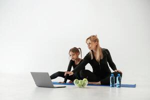 Laptop, apples, and bottles of water on the floor. Mother and little daughter are in fitness clothes on yoga mat photo