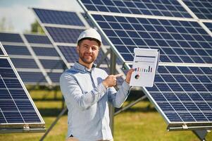 Pointing to the notepad with diagrams and,, graphs. Engineer with photovoltaic solar panels outdoors at daytime photo